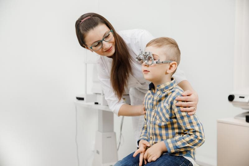 An optometrist fitting trial-frame glasses on a young boy during an eye exam.