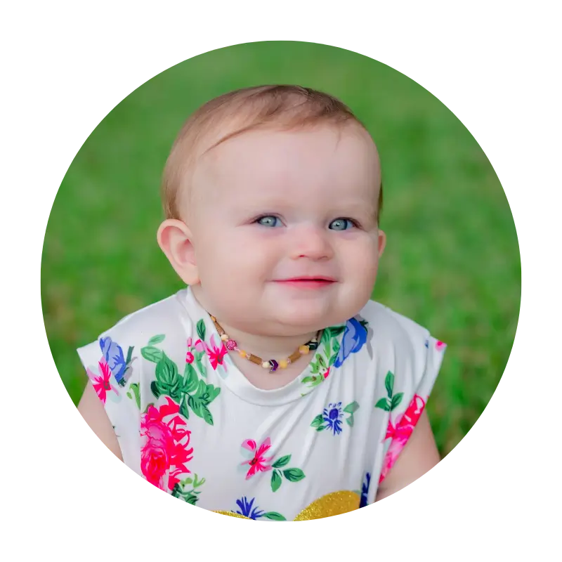 A baby in a floral dress is smiling in the grass.