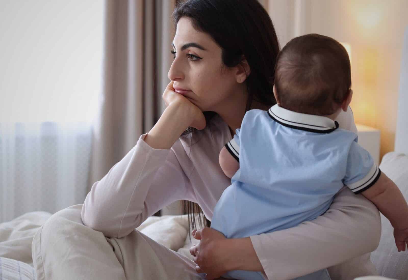 A mother sitting by a window holding her baby, looking thoughtful and tired.