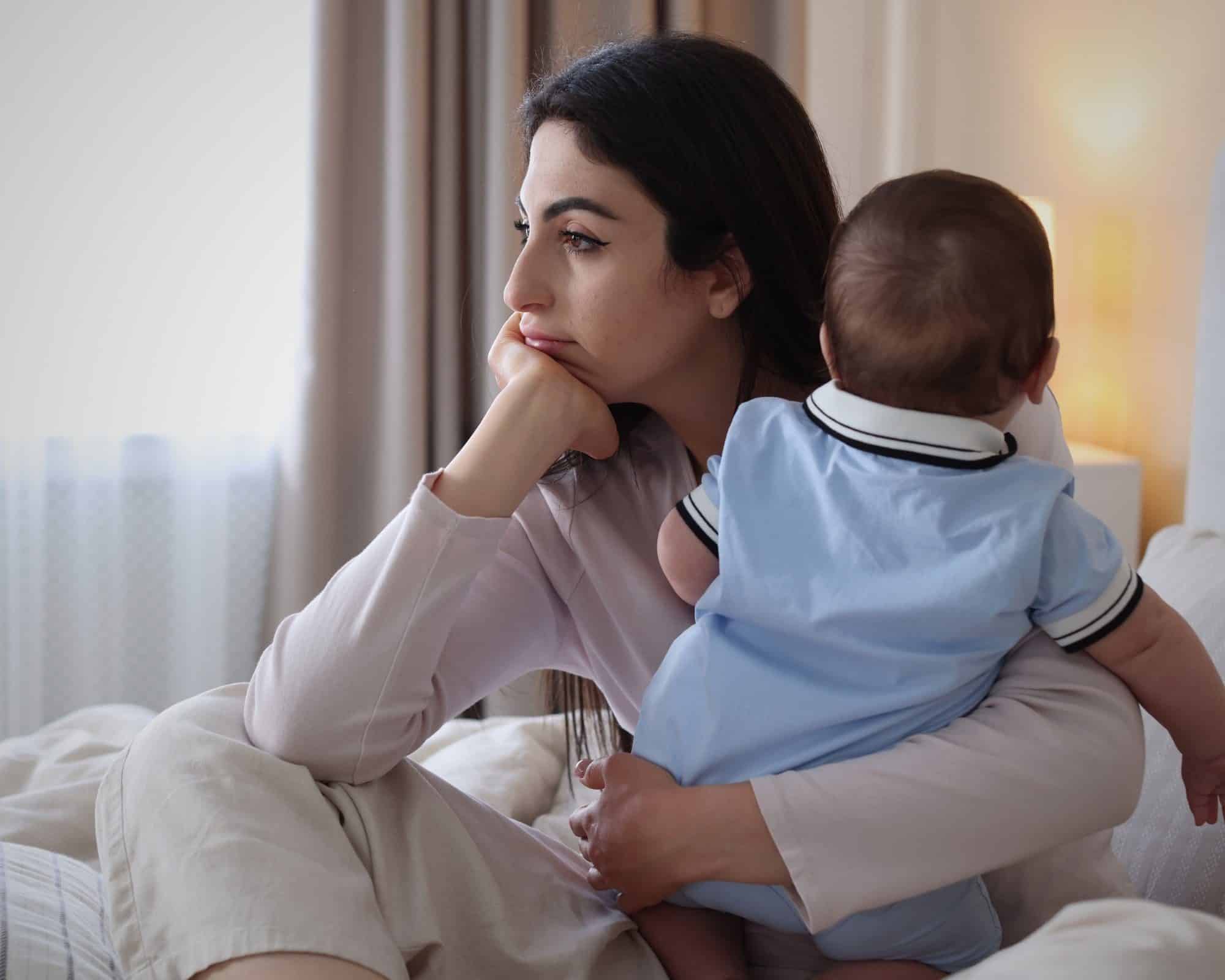 A mother sitting by a window holding her baby, looking thoughtful and tired.