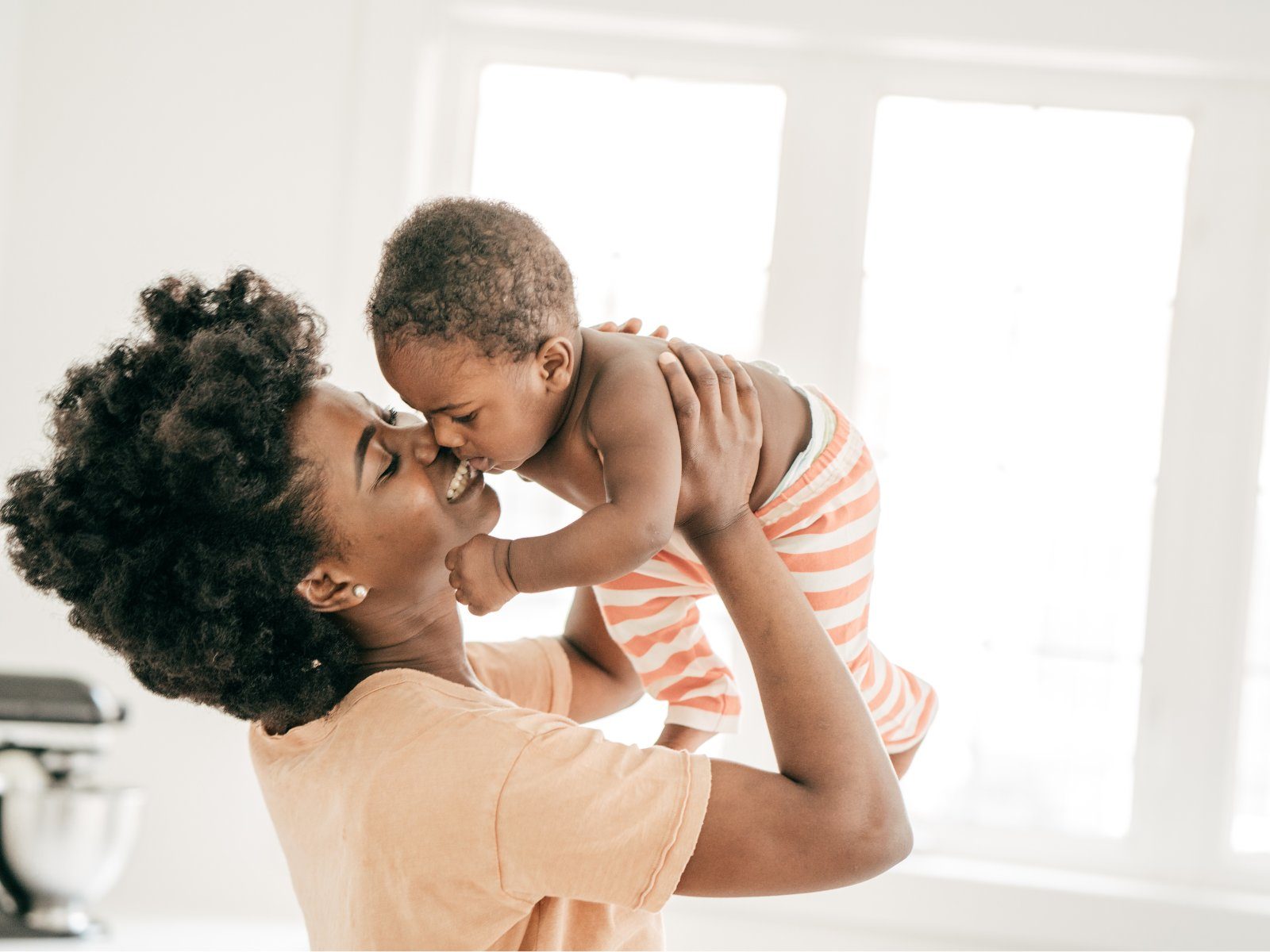 A smiling parent lifts and nuzzles their baby in a bright kitchen