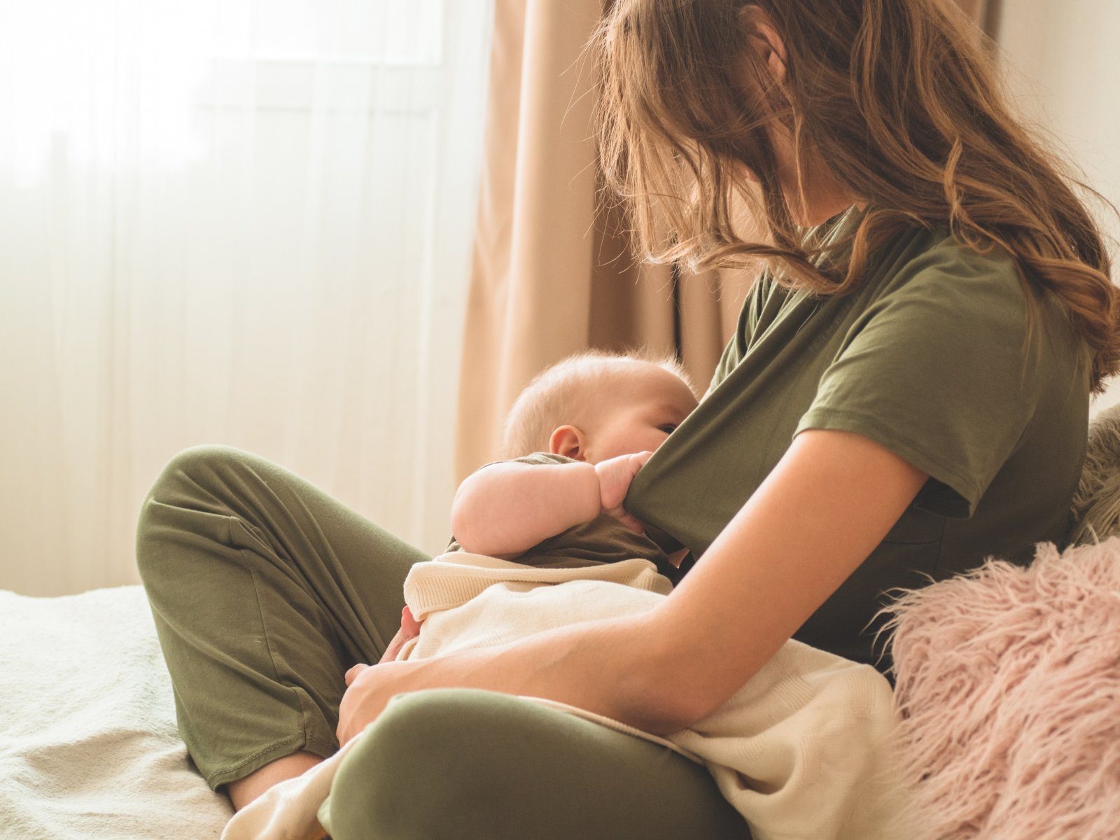 A parent breastfeeding their baby on a bed beside a sunlit window