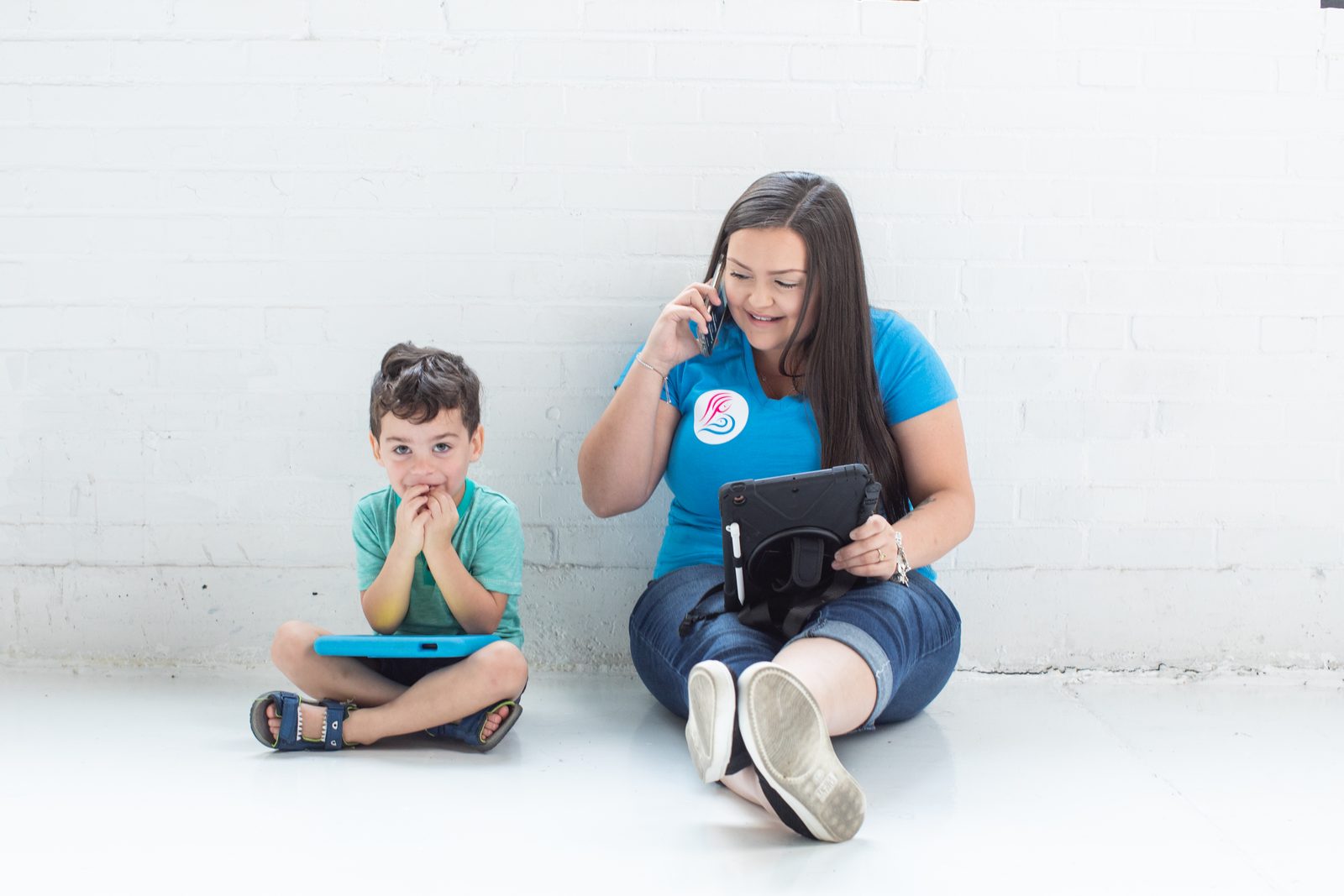 A smiling woman in a Bayou City Breastfeeding shirt talking on the phone while a young boy beside her plays on a tablet