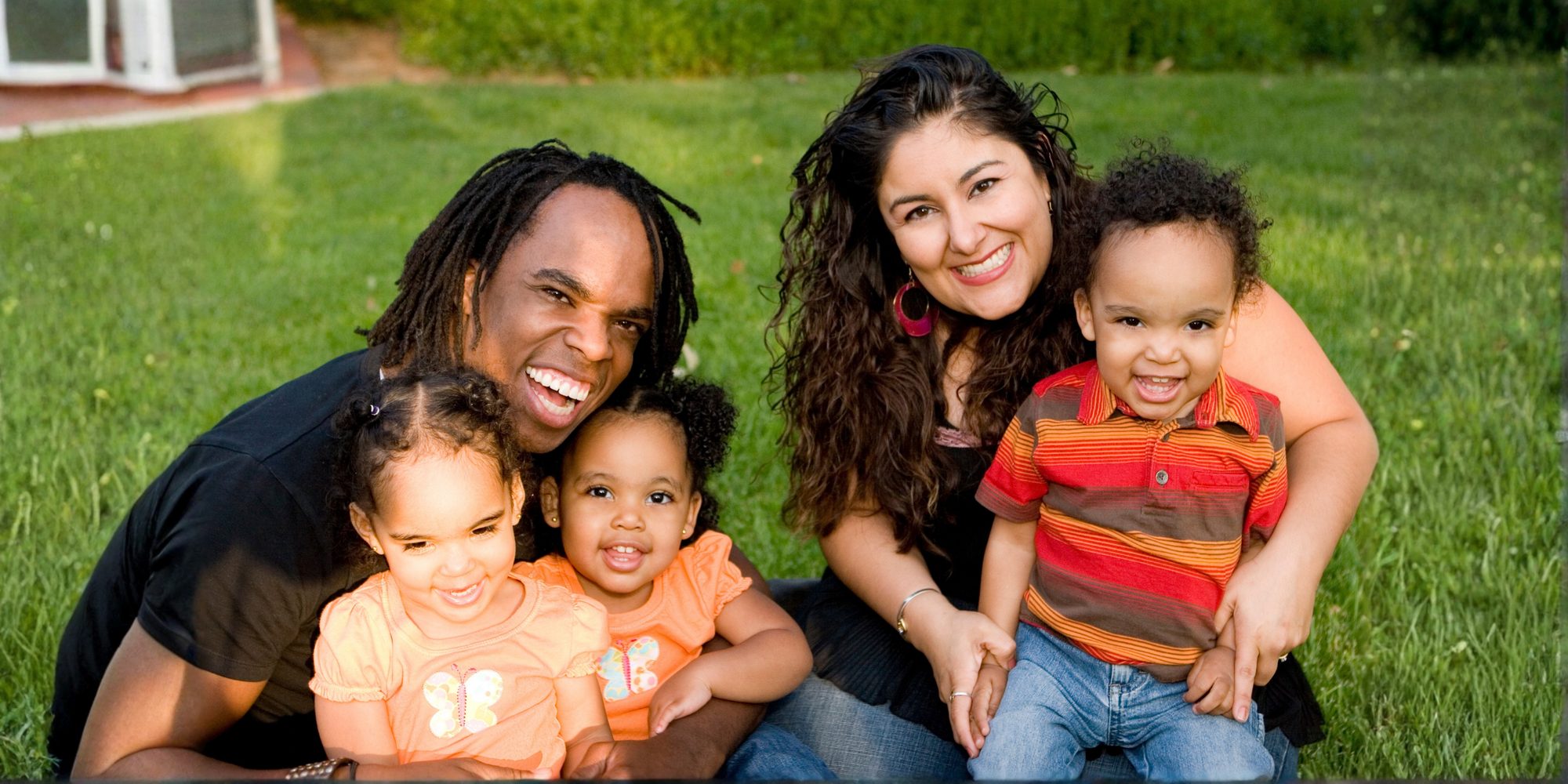 A smiling family of two parents and three young children sitting together on the grass