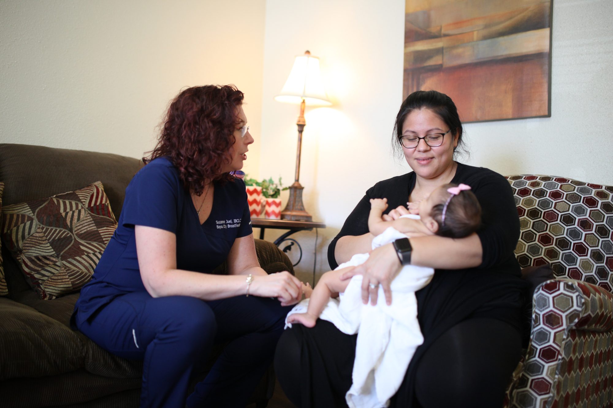 A parent smiling while breastfeeding their baby as an IBCLC sits alongside offering support during a lactation visit.