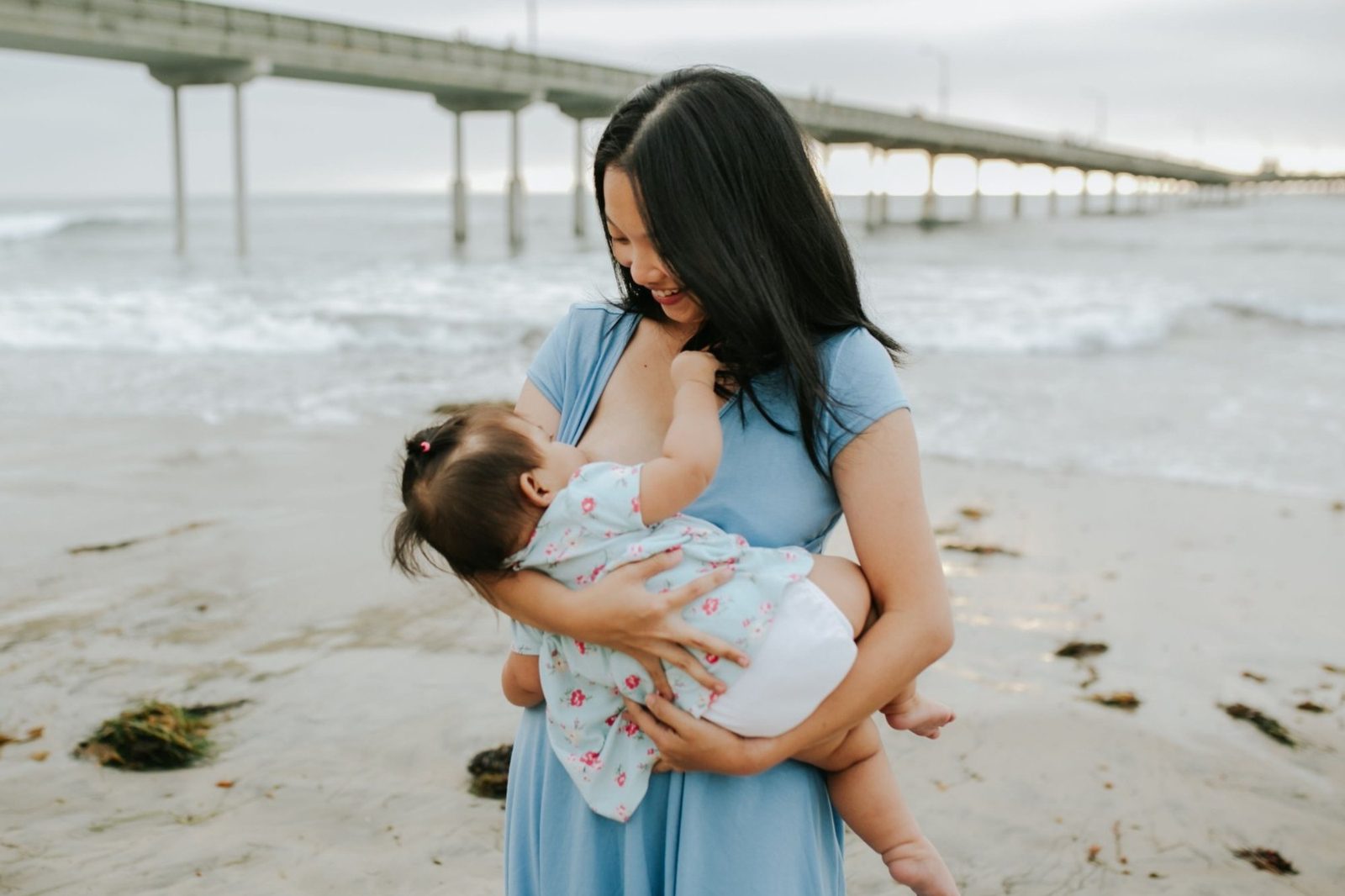 A smiling parent in a blue dress breastfeeding her toddler while holding her at the beach.