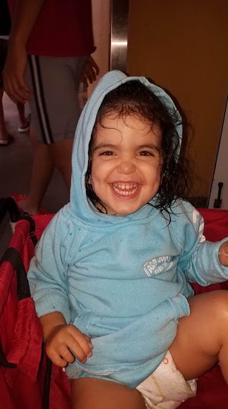 A smiling toddler in a blue outfit sitting in a car seat.