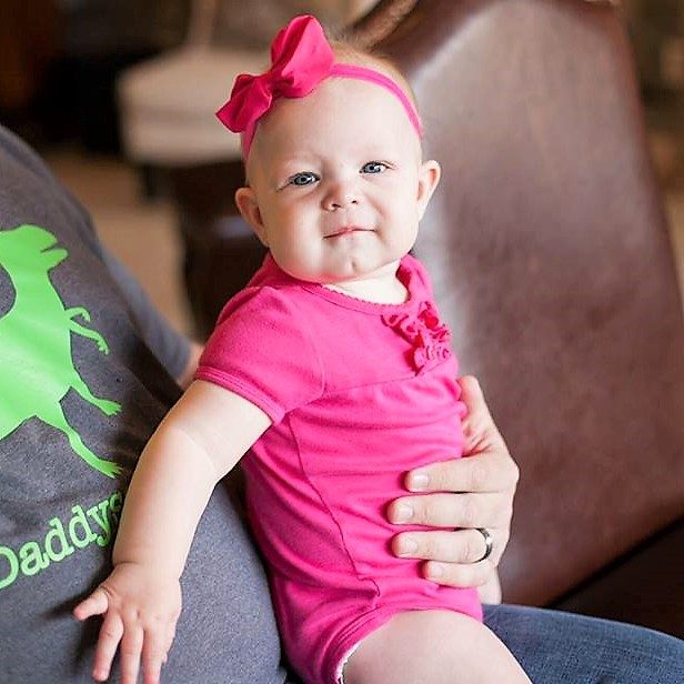 A baby in a pink bow and dress resting on a parent's lap.