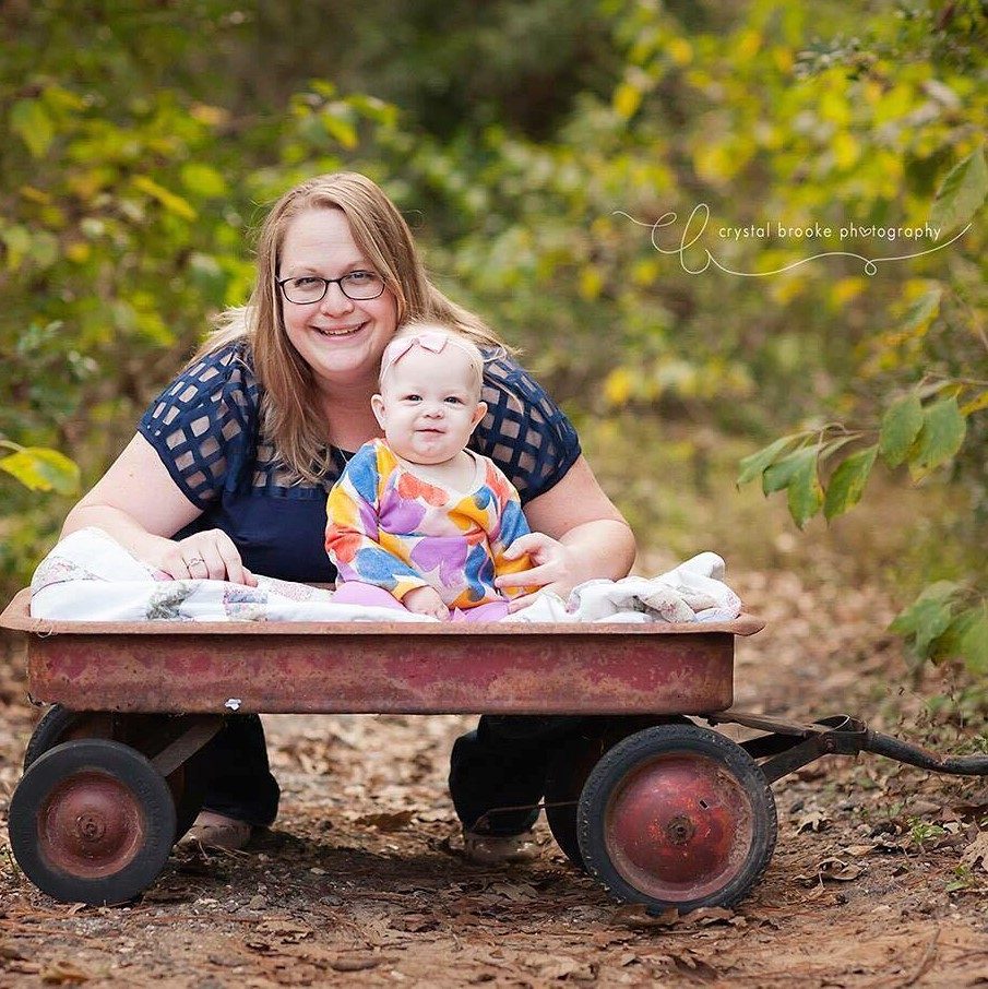 A mother kneeling with her baby in a small red wagon outdoors.