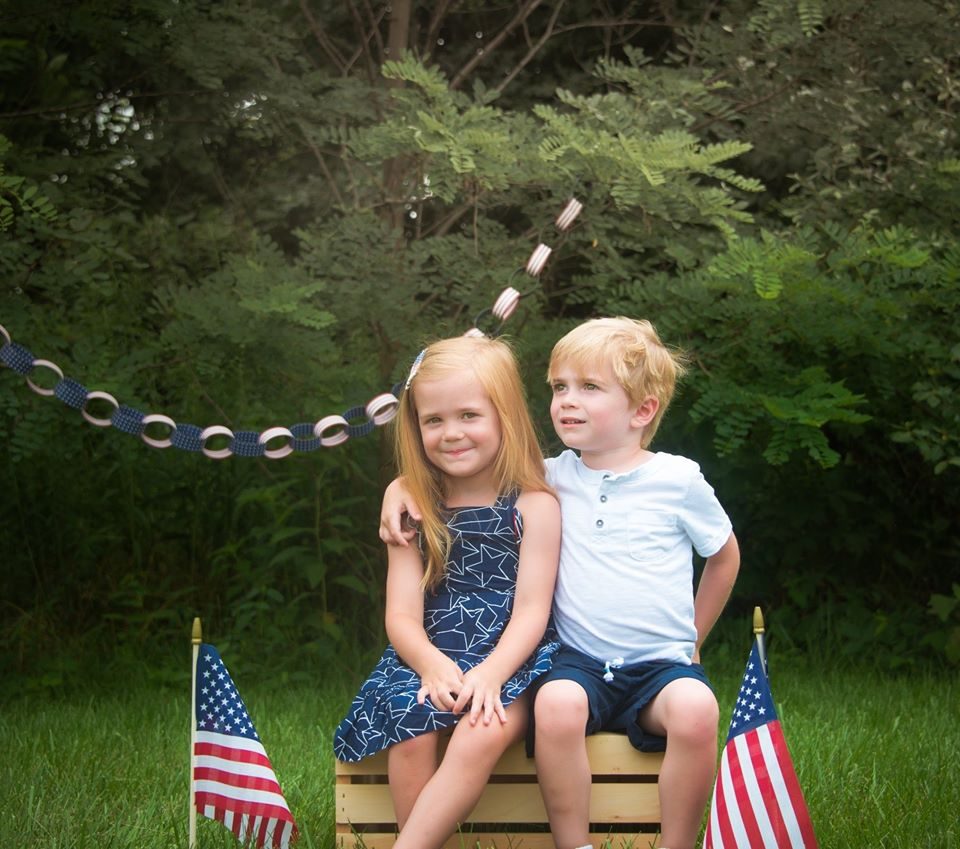 Two young siblings sitting together holding small flags outdoors.