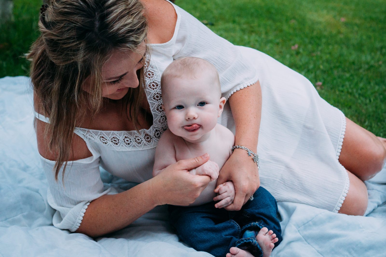 A mother kneeling on a blanket beside her smiling baby on the grass.