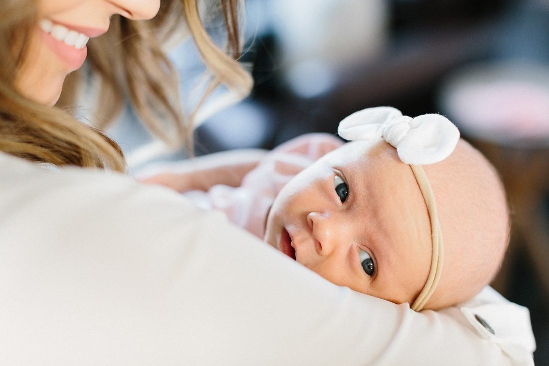 A wide-eyed newborn in a white bow headband.
