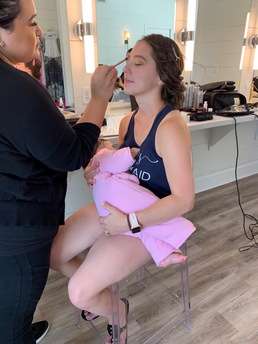A mother holding her baby while having her makeup done.