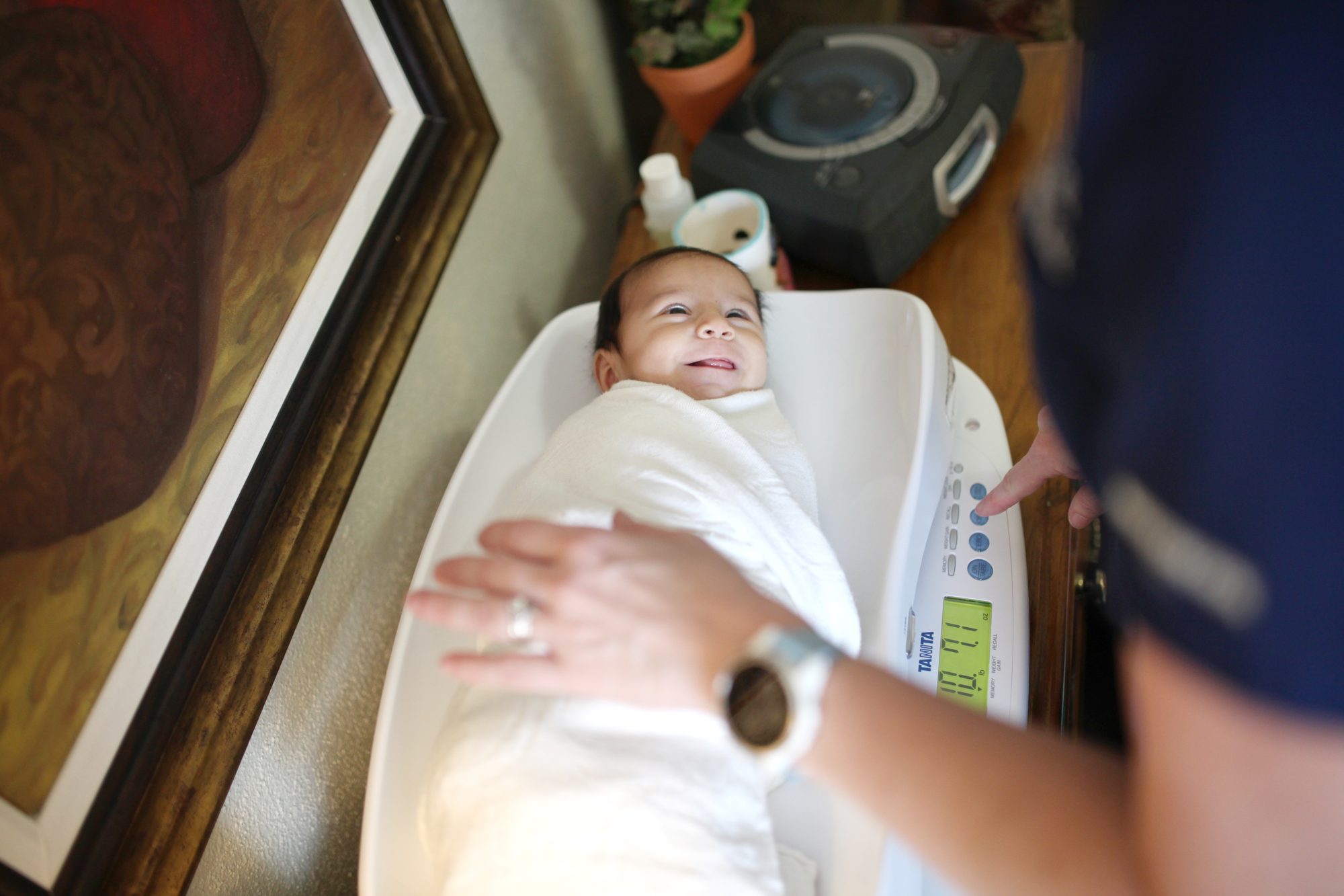 A smiling swaddled baby being weighed on a digital infant scale at home while an IBCLC steadies the baby and reads the scale