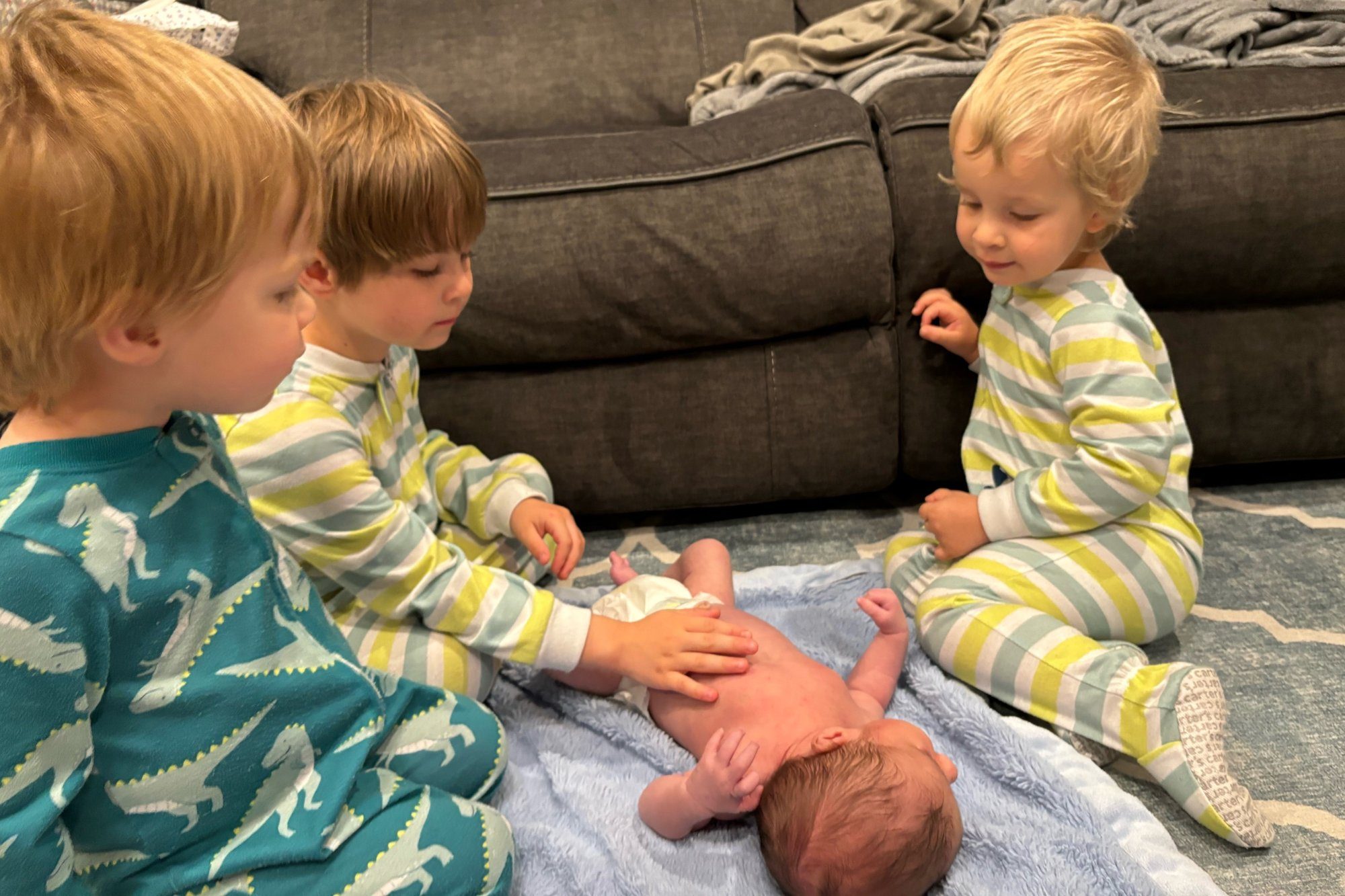 Three young siblings in pajamas sitting around their new baby on a soft blanket at home