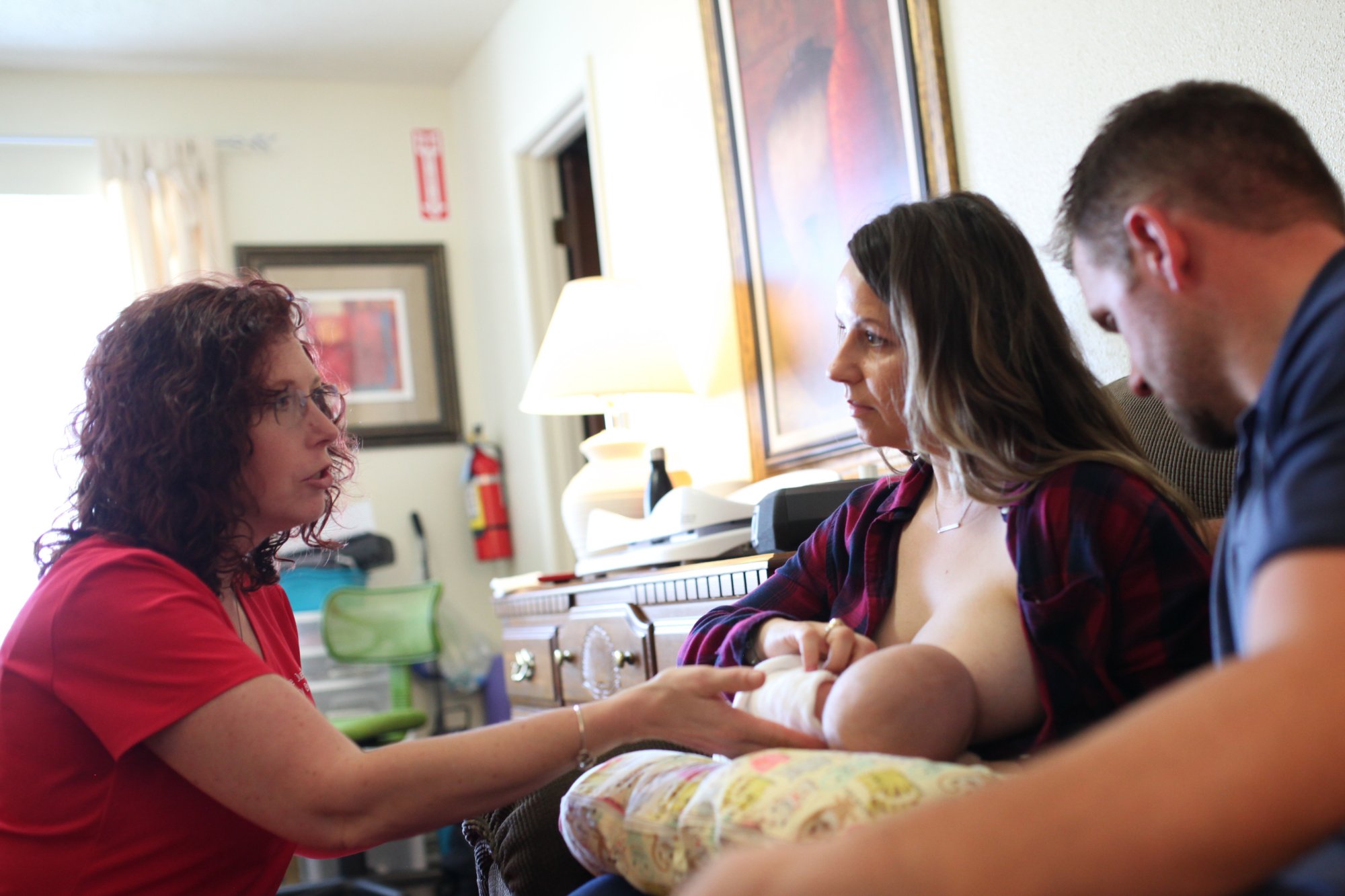 An IBCLC coaching a mother breastfeeding her baby while the father looks on during an in-home lactation consultation