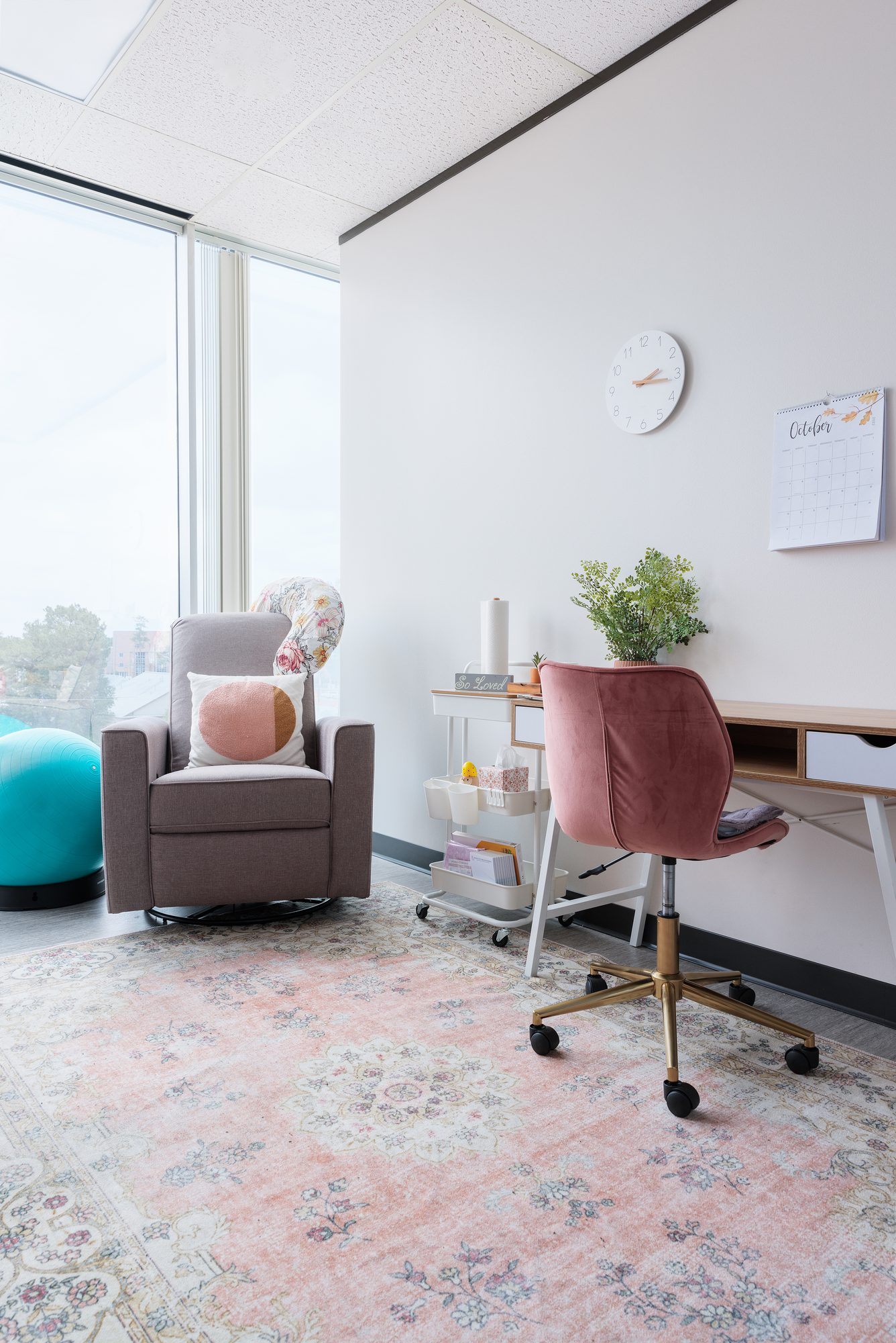 A sunny corner of a consult room with a glider, supply cart, and pink desk chair