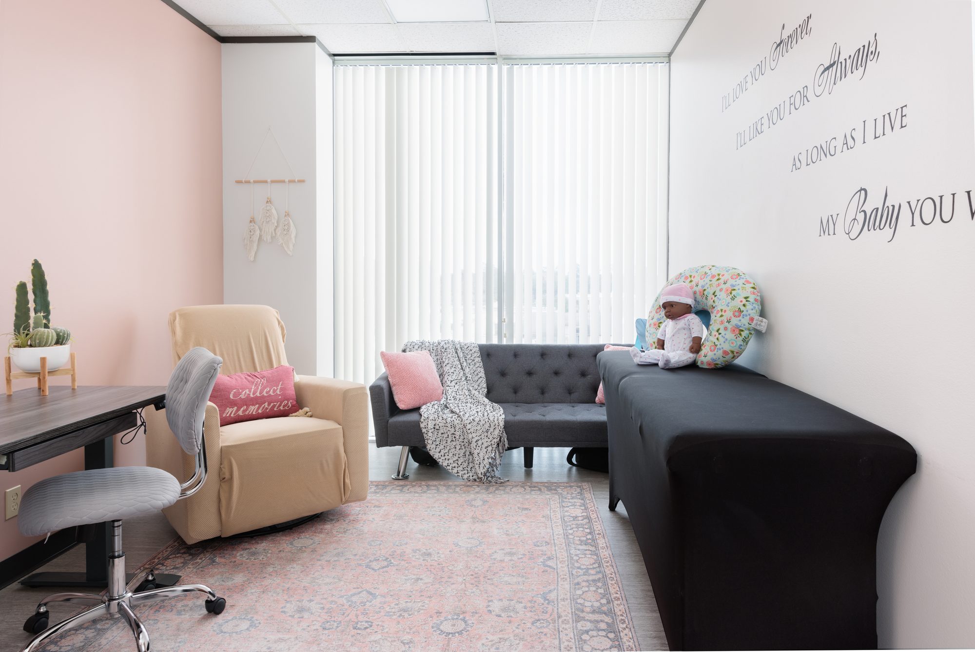A consult room with a soft pink accent wall, a cushioned glider, a grey sofa, and wall lettering that reads &lsquo;I&rsquo;ll love you forever&rsquo;