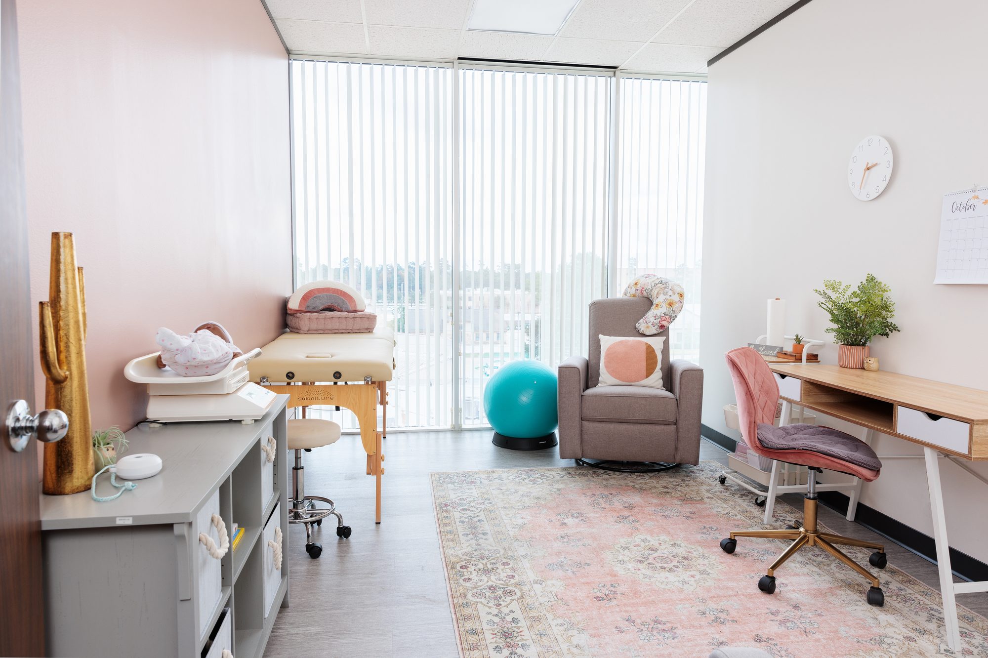 A bright corner consult room with floor-to-ceiling windows, a cushioned glider, an exercise ball, a massage table, and a pink desk