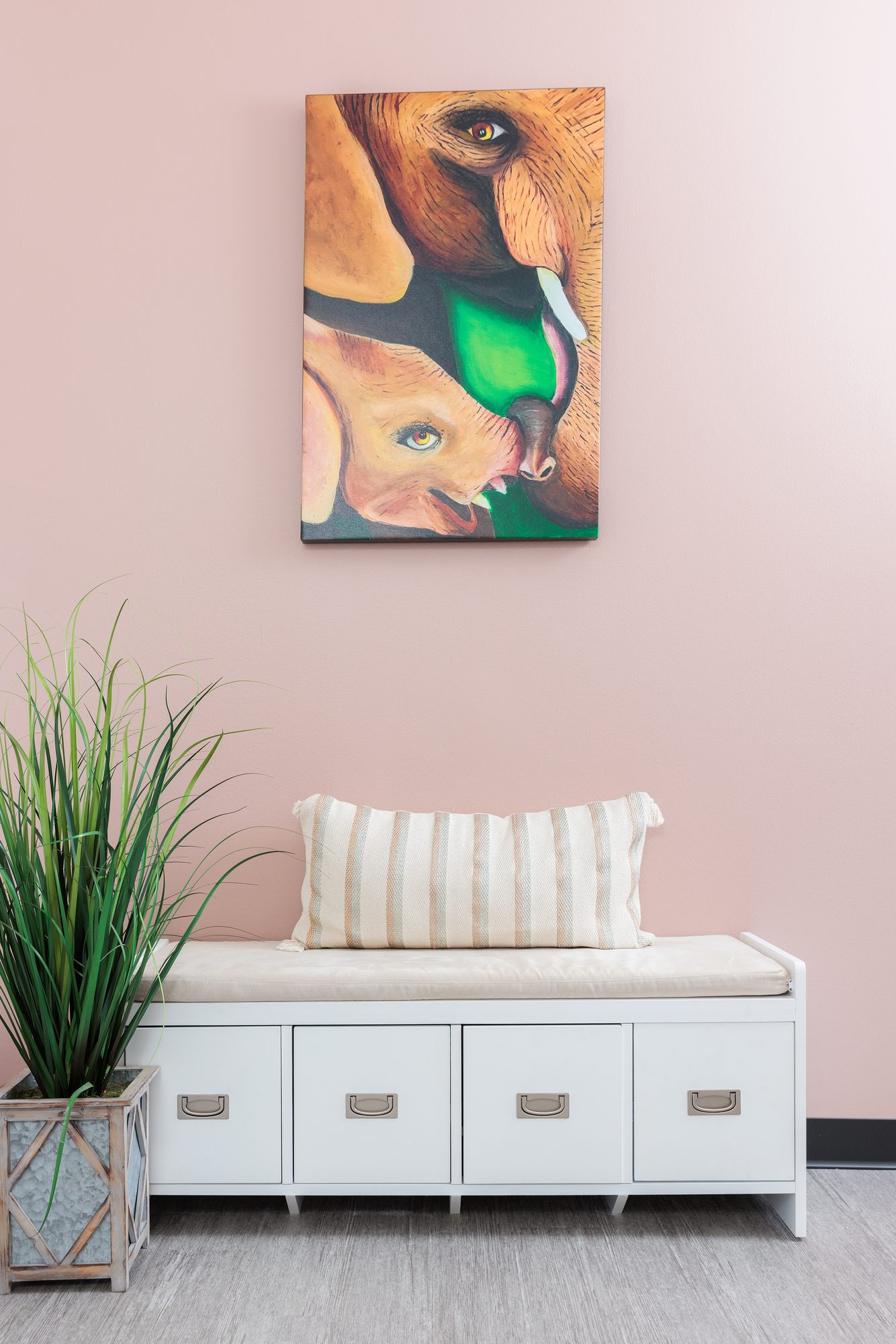A painting of an elephant and her calf above a cushioned storage bench and a tall grass plant