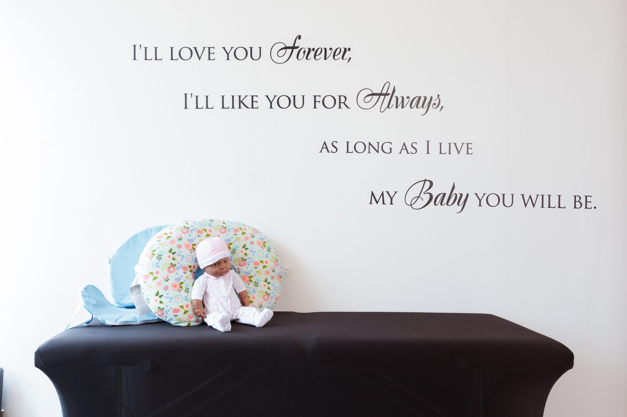 A padded table with a baby doll and nursing pillow beneath wall lettering
