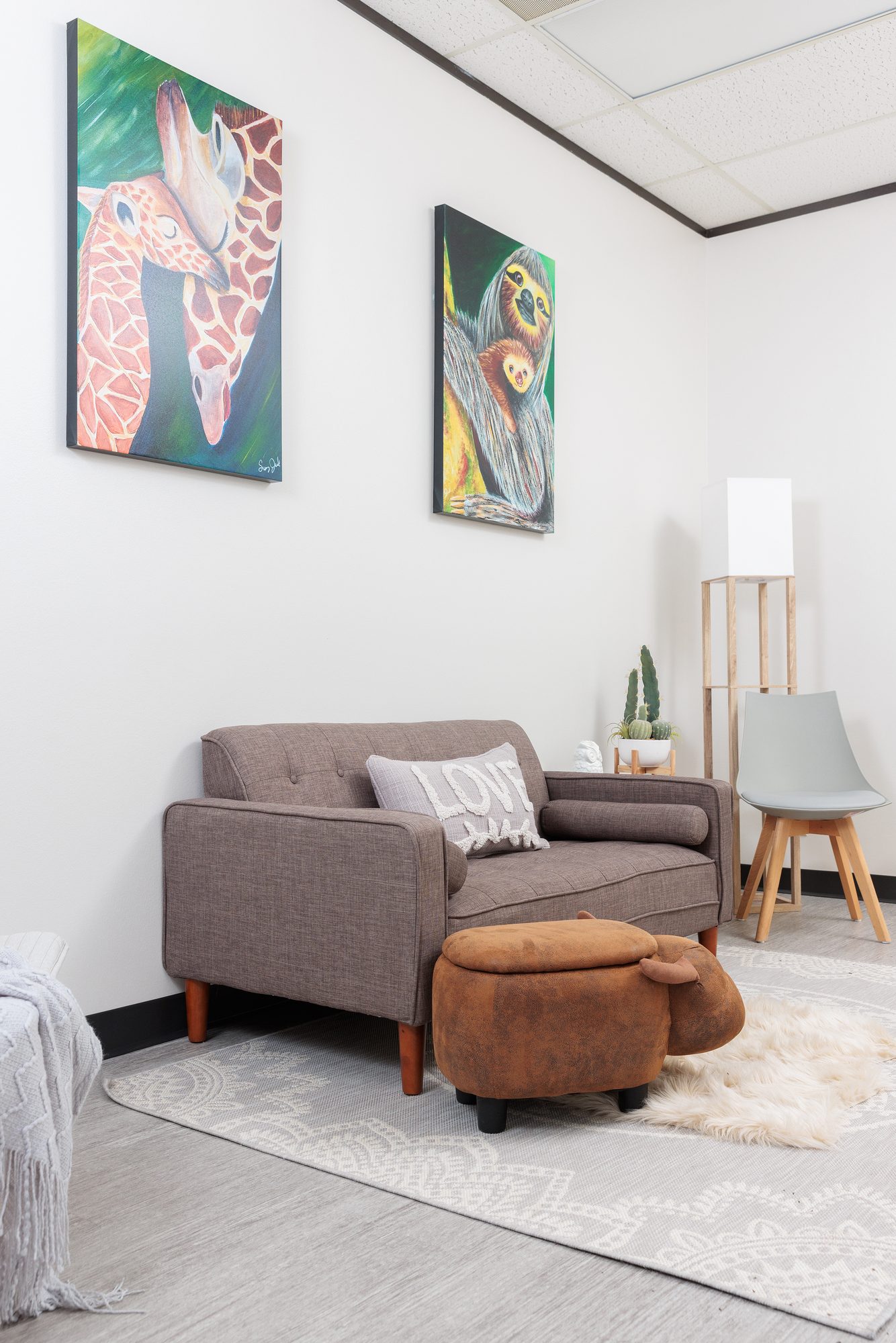 Paintings of a giraffe nuzzling its baby and a sloth holding its young, above a grey loveseat