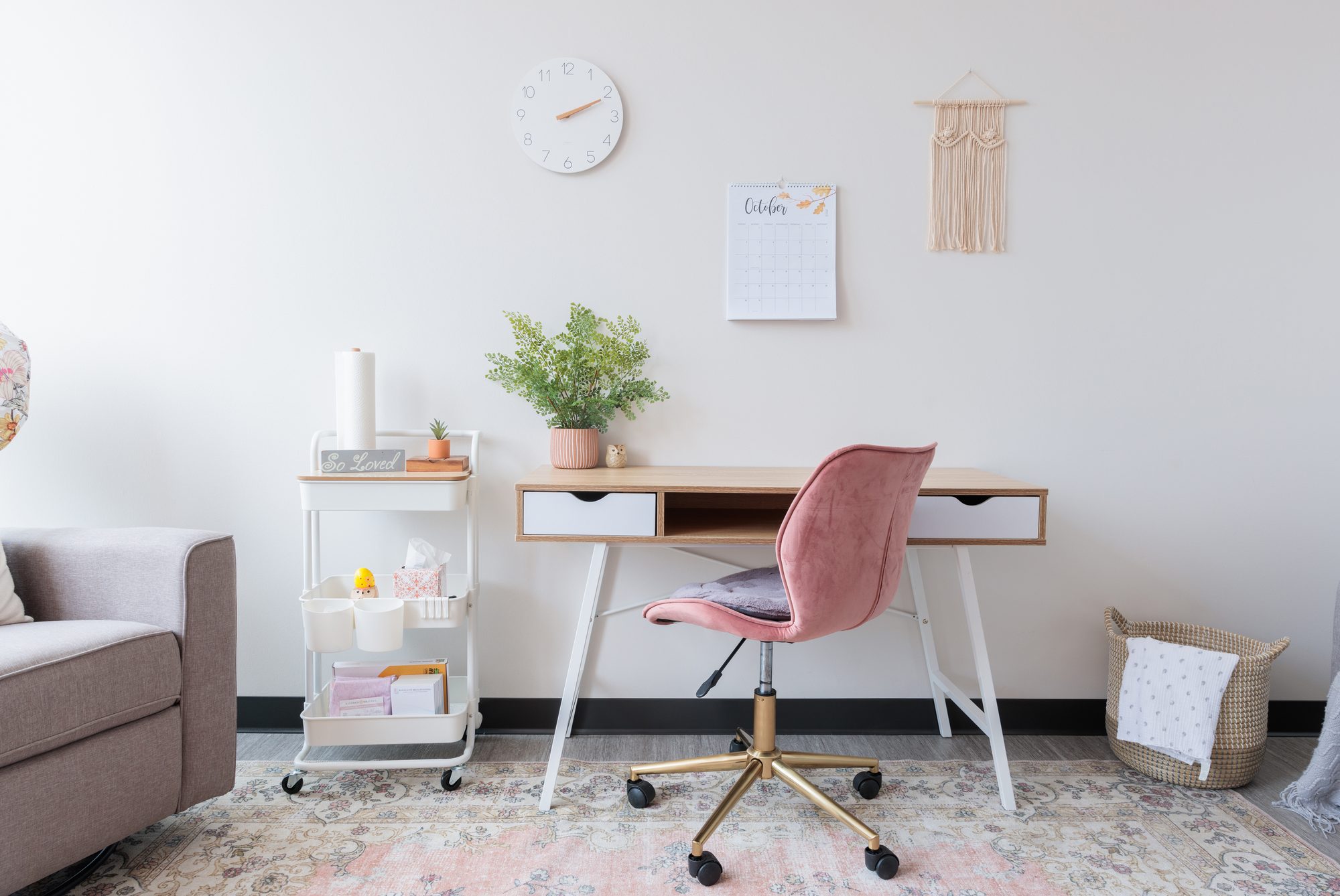 A tidy desk nook with a pink velvet chair, a wall calendar, a clock, a macrame hanging, and a stocked supply cart