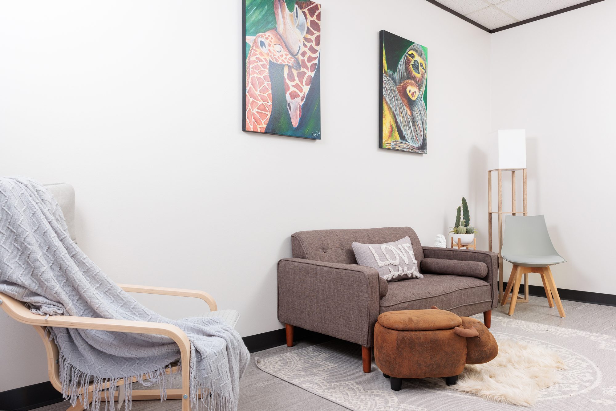 A grey loveseat with a &lsquo;LOVE&rsquo; pillow beneath giraffe and sloth paintings, with a hippo ottoman on a sheepskin rug