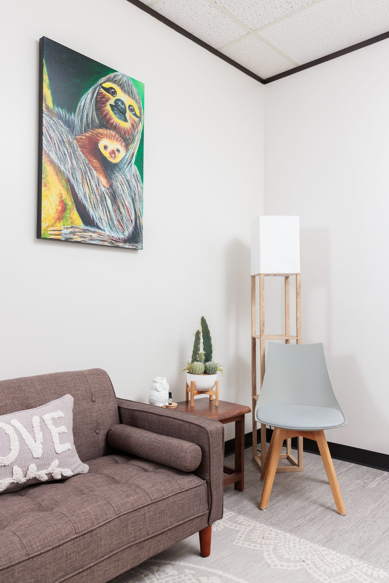 A sloth-and-baby painting in a cozy corner with a floor lamp and a small cactus