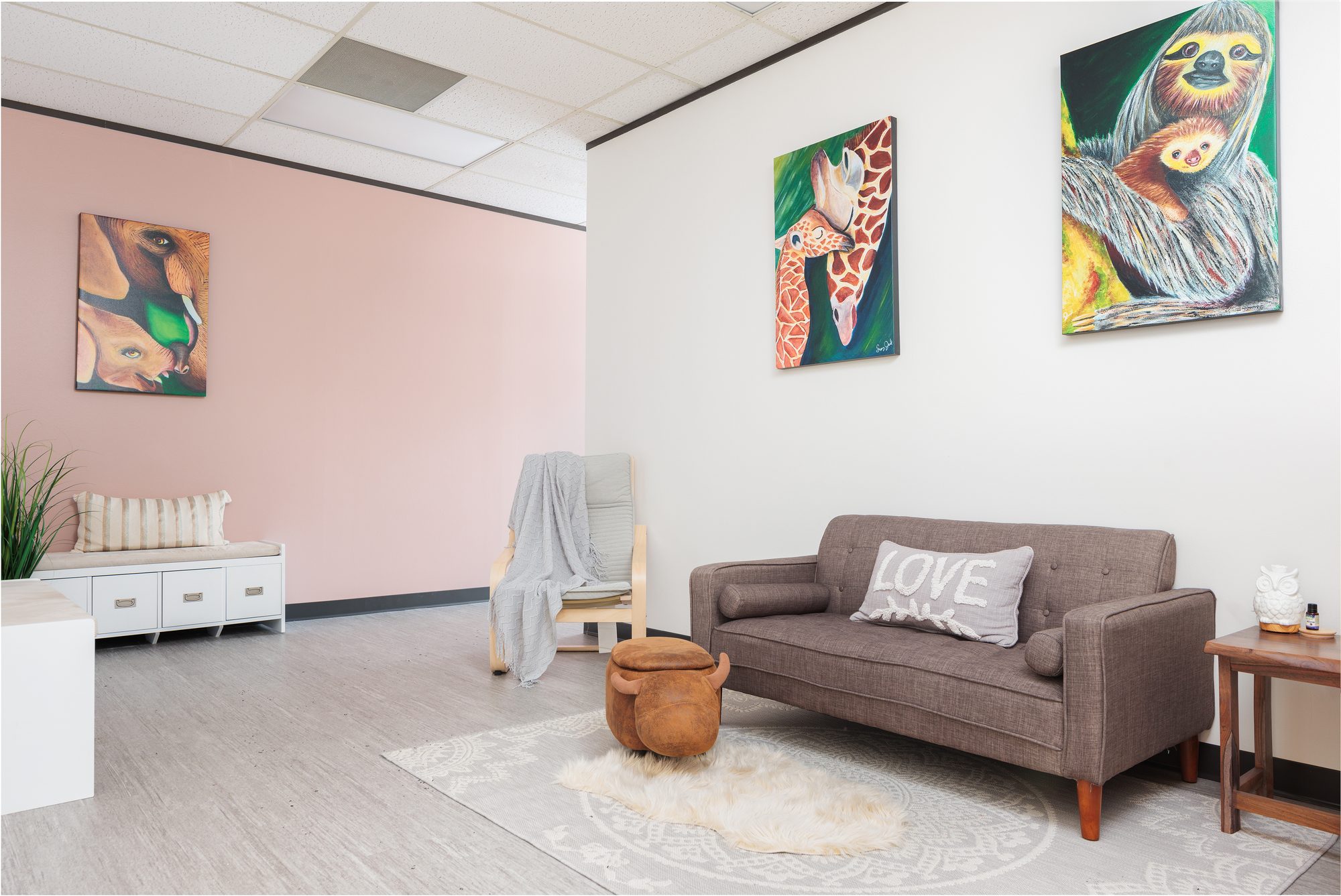 A waiting area with a pink accent wall, a grey sofa with a &lsquo;LOVE&rsquo; pillow, a storage bench, and colorful animal paintings
